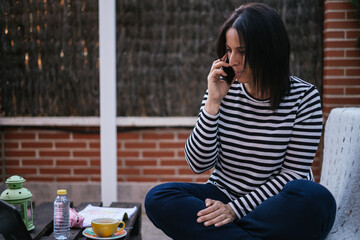 Brunette woman talking on the cellphone smiles in the garden of her house while teleworking with her laptop. She is wearing dark jeans and a blue and white striped sweater