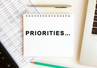 Notepad with text PRIORITIES on a white background, near laptop, calculator and office supplies. Business concept.