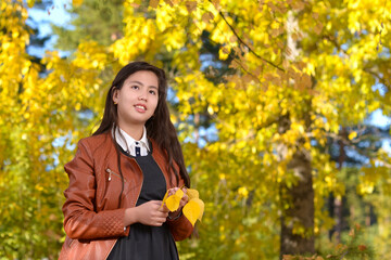 A girl in a brown leather jacket holds yellow leaves. Portrait of a beautiful young girl with long black hair against the background of the autumn forest.
