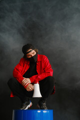 An emotional young man 25-30 years old in a black protective mask, a cap and a red jacket holds a megaphone against a dark background. 