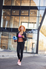 Young european redhead woman in sportive clothes with phone and mat in hands outdoors