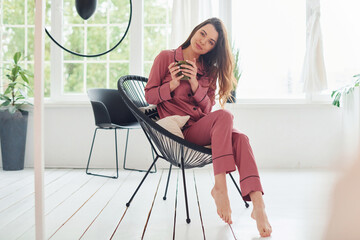 Cheerful young woman in pajamas sitting on the chair indoors at daytime