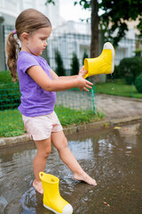  Cute girl jumping in puddles for walking near the house on  summer day. Rubber boots full of water. Children are having fun outside after rain. Outdoor activities in bad weather