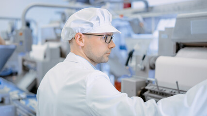 Naklejka premium Young Male Food Conveyor Belt Employee Works at a Dumpling Factory. He is Pushing a Button. He Wears White Sanitary Hat and Work Robe.