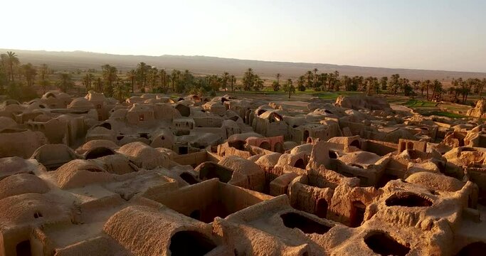 Fly over ruins of mud houses of a desert village in Iran beside saffron fields and date palms tree gardens in sunset