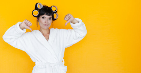 young beautiful woman housewife in a white robe. curlers on the head. Yellow background. looks at the photographer with her thumbs down.