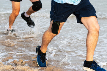 Fototapeta premium Legs of runners running in water on the edge of beach with cell phone sticking out of shorts