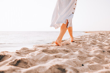 Cropped image of young barefoot woman in a dress walking alone on the beach in the sunset. Face is...