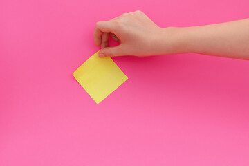 Woman's hand glues yellow sticker or sticky post-it notes on pink background.
