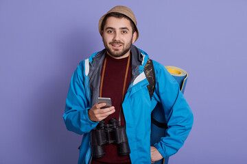 Smiling traveler man in cap and with backpack isolated on blue background. Tourist traveling on weekend getaway, being in good mood, holding smart phone. Tourism, discovering, hiking.