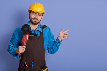 Bearded Caucasian builder with grinder in hands posing isolated over blue background and pointing index finger aside at copy space for advertisement