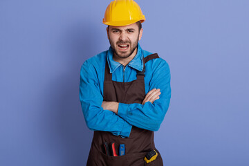 Annoyed emotional workman wearing yellow protective construction helmet, shirt and brown apron, has...