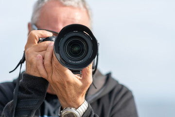 A middle-aged man, in his 50th, takes a picture with a modern mirrorless camera. The camera covers his face. Blue hazy sky in the background