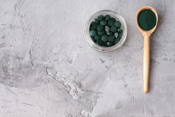 Green chlorella powder in wooden spoon and green chlorella tablets in small glass jar  on gray concrete background. Chlorella is a single-celled green algae, it is used to make nutritional supplements