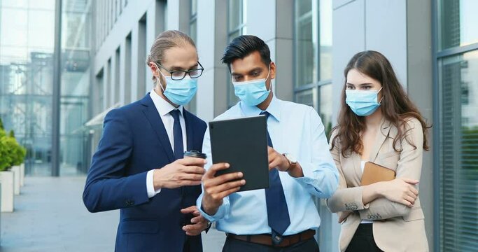 Mixed-races young males and female business employees in medical masks and with coffee-to-go standing outdoor and watching at tablet device. Multiethnic businessmen and Caucasian woman using gadget.