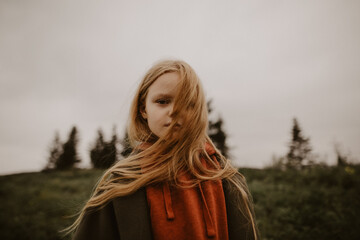 a little girl with blond hair looks seriously into the distance against the background of woods and...