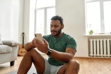  Distraction. Young man using smartphone app while having morning workout at home. Freshman relaxing after training. Online personal trainer on mobile phone © Svitlana