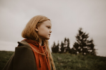 a little girl with blond hair looks seriously into the distance against the background of woods 