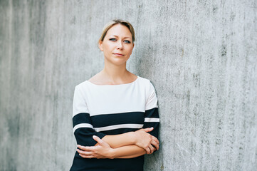 Outdoor portrait of middle age woman, leaning on grey wall, arms crossed