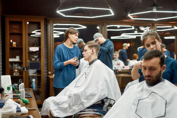 People in barbershop. Young professional barber girl, female hairdresser working with hair clipper, serving young handsome guy sitting in barber shop chair