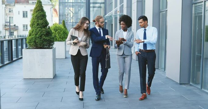Mixed-races young cheerful people in business style walking the street , laughing and talking. Multiethnic business white-collars males and females smiling and going to work in morning. Men and women.