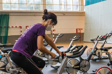 Young brunette caucasian girl with face mask on a stationary bike in an empty gym due to the low capacity in the new normal, social distance, covi-19, coronavirus, pandemic