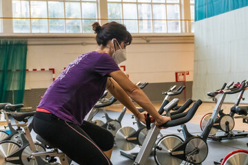 Young brunette caucasian girl with face mask on a stationary bike in an empty gym due to the low capacity in the new normal, social distance, covi-19, coronavirus, pandemic