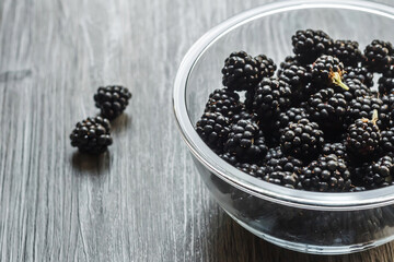 Berries of fresh ripe blackberries in a glass bowl on a gray wooden background. Healthy summer berries. Healthy vegetarian food. Background with blackberries and free space.