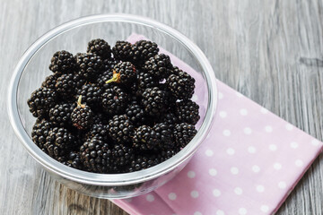 Fresh ripe blackberries in a glass bowl on a gray wooden background with a pink cloth. Healthy summer berries. Healthy vegetarian food. Background with blackberries and free space.