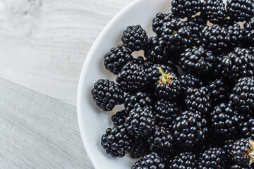 Berries of fresh ripe blackberries in a white plate on a gray wooden background. Healthy summer berries. Healthy vegetarian food. Background with blackberries and free space.