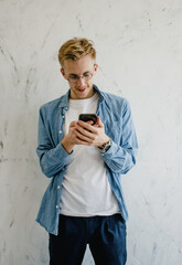 A young hipster guy in round glasses and a blue shirt checks the mail on his phone standing on a white background