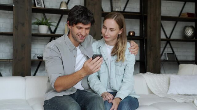 Сouple sitting on sofa at home. Man and woman relaxing on couch with smartphone in living room.