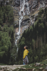 Naklejka na ściany i meble Young brunette girl in yellow sweatshirt closed eyes & feels freedom on background of Rothbach waterfall, pine forest & rock mountain cliff. Schonau am Konigssee. Bavaria. Germany
