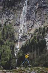 Obraz premium Young brunette girl in yellow sweatshirt jumps on background of Rothbach waterfall, pine forest & rock mountain cliff. Schonau am Konigssee. Bavaria. Germany