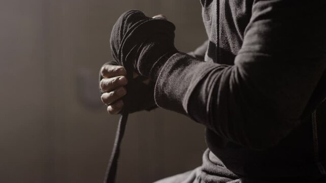 Boxer, Muay Thai fighter putting on hand wraps while sitting on a bench in a musty locker room prior to a fight close up in slow motion