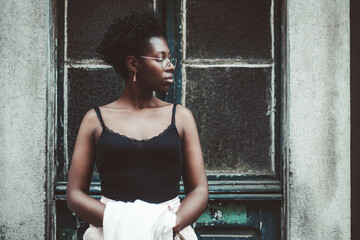 Portrait of the young black female in a tank top and spectacles looking aside while standing in front of an old antique wooden door with glass inserts and holding a white trench in her hands