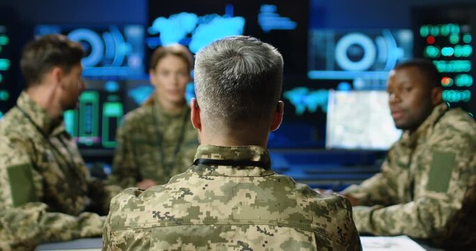 Rear of general with mixed-races militarian council sitting at table, deciding war or defense tactics in dark monitoring room. Back view of multiethnic people, officers talking, discussing terrorism.