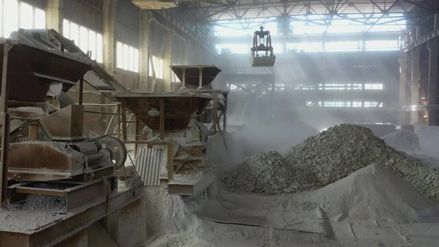 production workshop, crushing stones, huge bucket pick up stone blocks and lifts  into powerful unit. fractions and sorting on carpet line. rays of sun through dusty workshop, chemical industry grind
