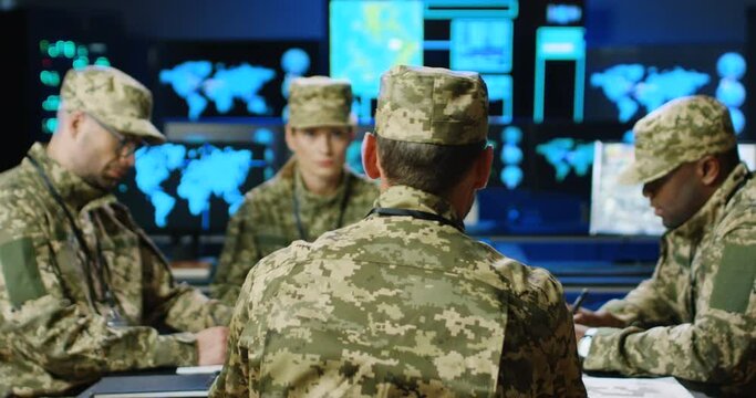 Rear of general giving orders mixed-races militarian council sitting at table, deciding war or defense tactics in dark monitoring room. Back view of multiethnic officers talking, discussing terrorism.