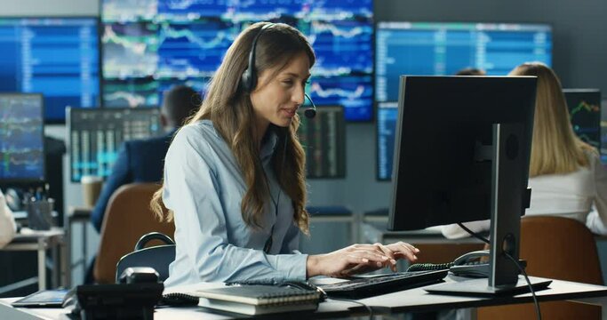 Young Caucasian pretty woman trader in headset working at monitor of computer in trading office and talking with client. Female broker typing on keyboard and speaking with customer. Sales call center.
