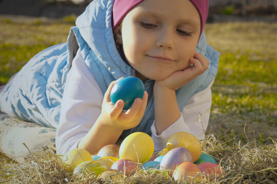 Cute little girl lying on green lawn and plays with colored easter eggs