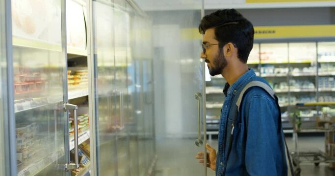 Handsome young Hindu man in glasses and blue jeans shirt standing in supermarket, opening refrigerator and taking out butter, yoghurt or cheese. Guy student reading sticker and putting back.