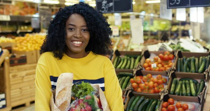 Portrait of happy beautiful African American woman with curly hair and in yellow sweater standing in grocery shop and holding packet with food. Pretty girl smiling to camera with bag of healthy food.