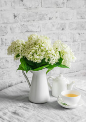 Bunch of hydrangeas in a vintage jug, cup of green tea, teapot on the table. Cozy home still life