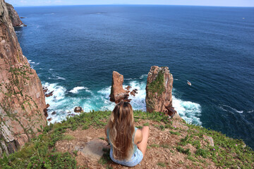 The girl sits on a cliff and looks at the rocks in the sea