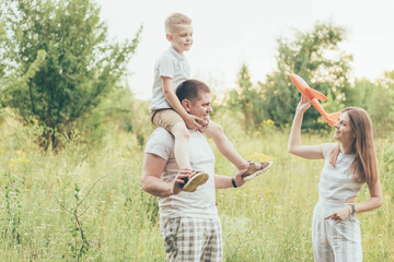A family plays in nature, a boy sits on his father's neck and mom holds a toy plane in her hands