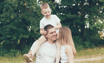 Family resting in nature, dad keeps the boy around his neck and mom kisses dad and smiles