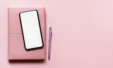 Top view of blank screen smartphone with notebook and pen on pink leather top table, copy space.