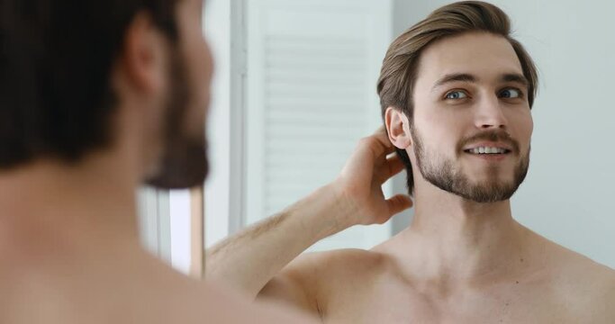 Smiling shirtless caucasian man looking at mirror, combing hair with fingers, getting ready for new day in bathroom. Head shot close up happy young stylish guy enjoying morning beauty routine indoors.