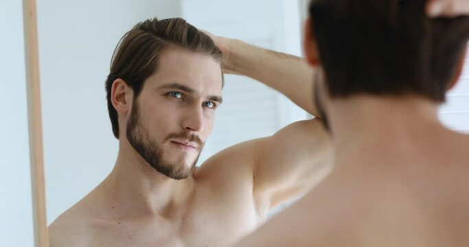 Young serious handsome european man looking at mirror, combing styling hair with fingers, assessing hair condition, worrying about dandruff or thinking of visiting barbershop and haircut service.